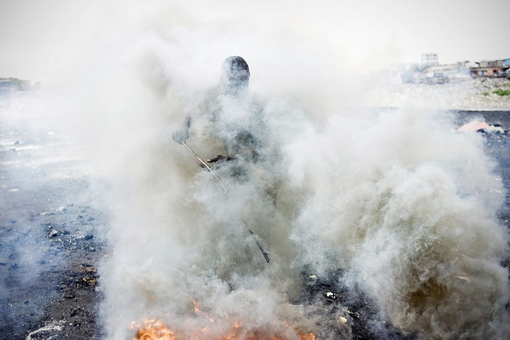 In eine dichte Rauchwolke gehüllt stochert ein Mann mit einem dünnen Stab in einem Feuer.
