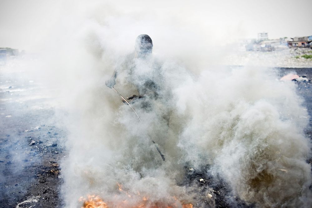 Shrouded in a thick cloud of smoke, a man pokes at a fire with a thin stick.