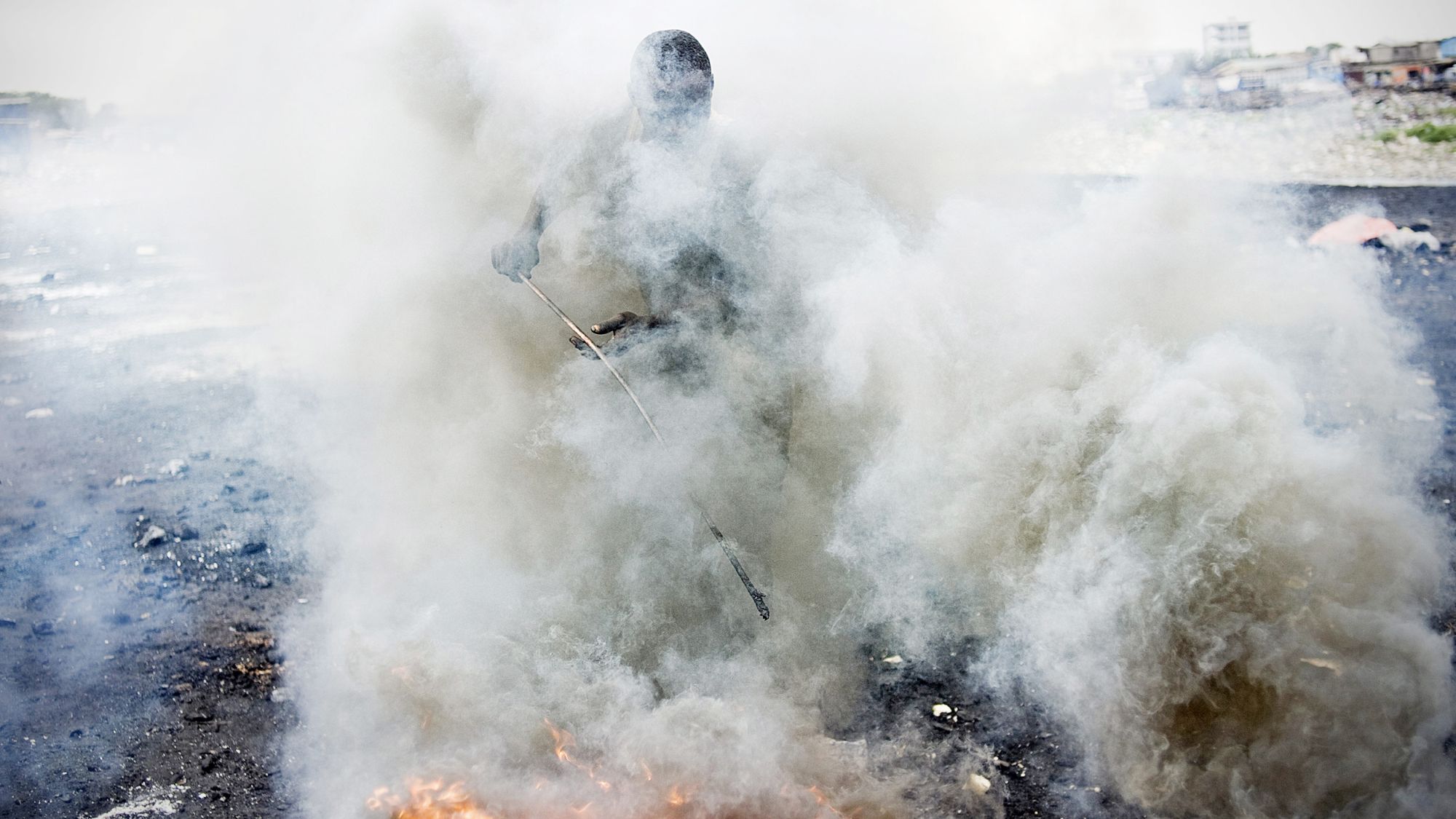 Shrouded in a thick cloud of smoke, a man pokes at a fire with a thin stick.