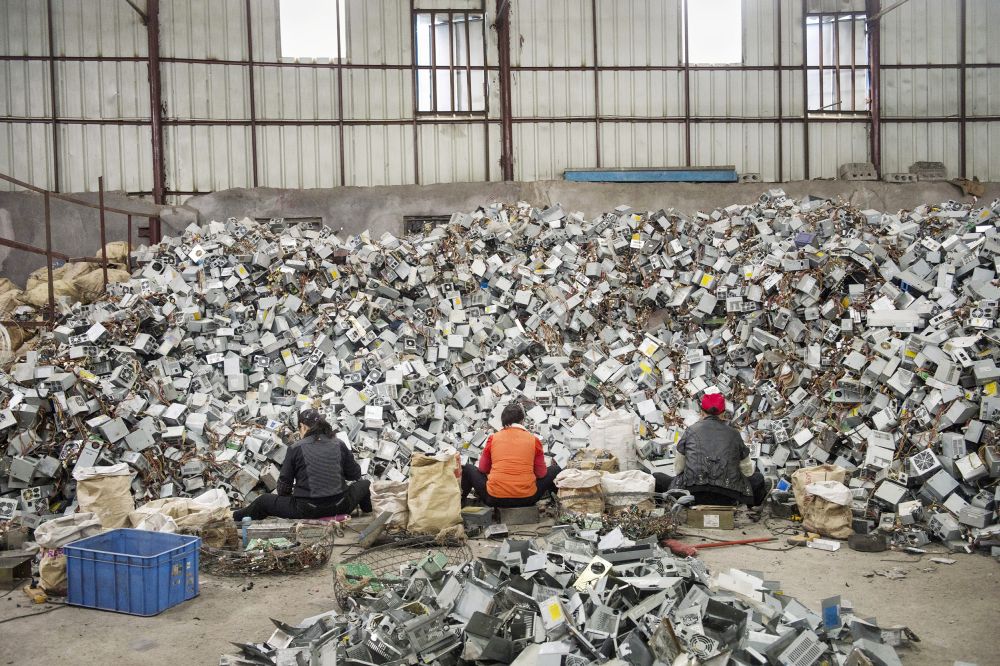 Three women sit with their backs to the viewer in front of a large pile of computer casings.