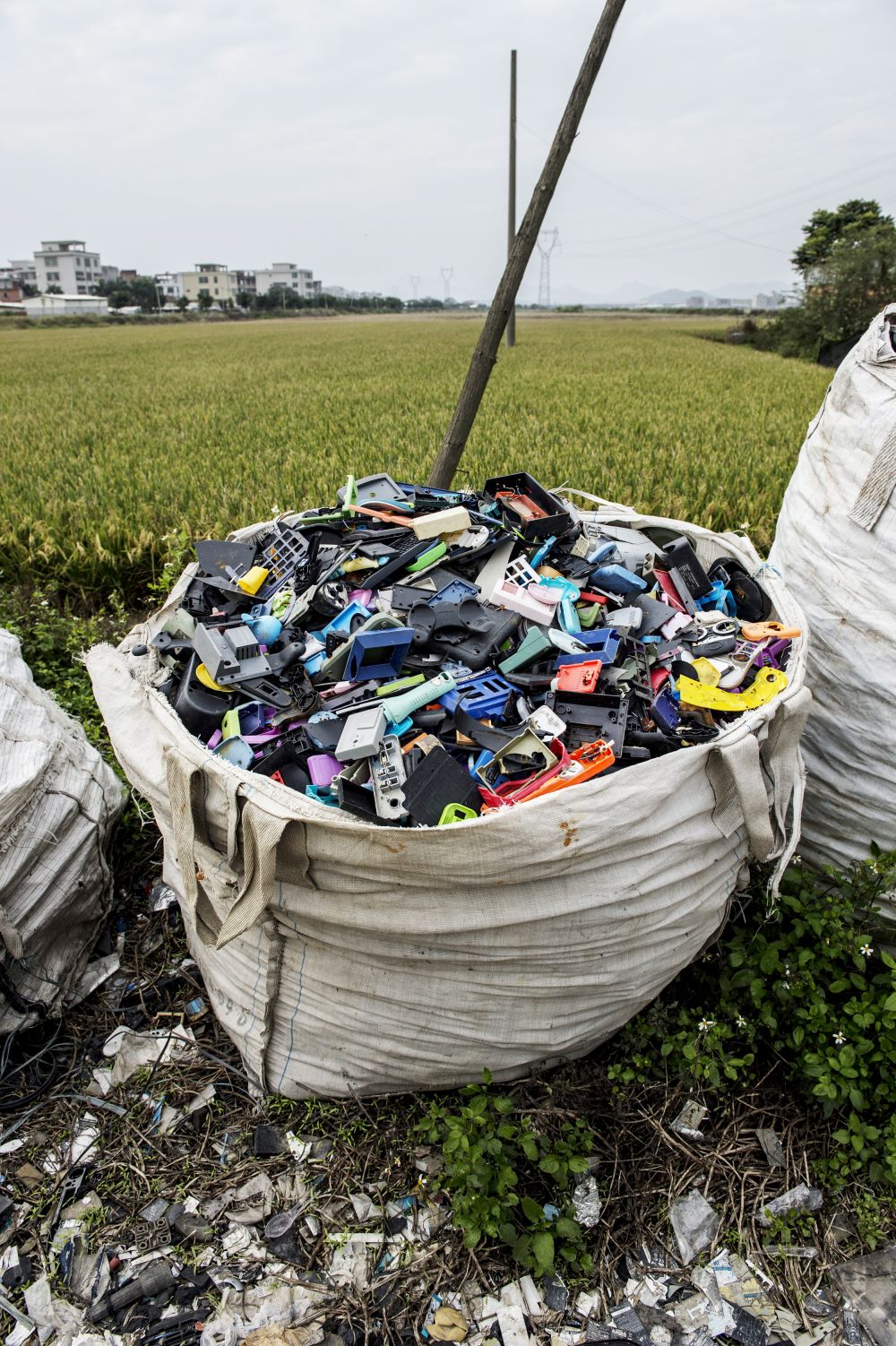A large plastic bag stands in front of a field and is filled with plastic waste, including individual parts of gaming controllers.