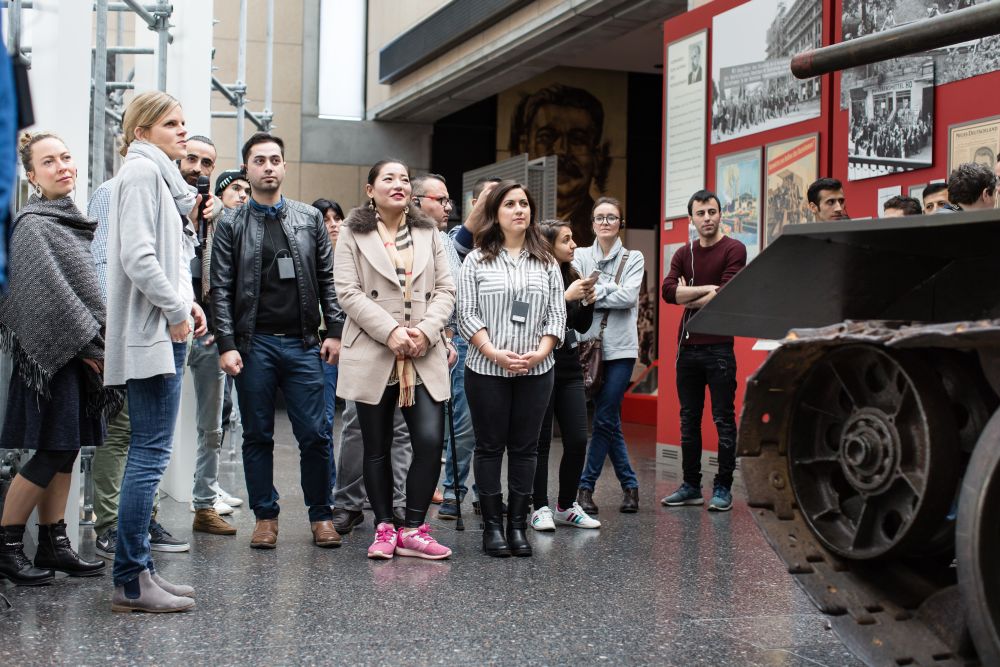 Eine Gruppe junger Erwachsener steht in der Ausstellung dem Betrachter zugewandt, recht im Bild der vordere Teil eines Panzers