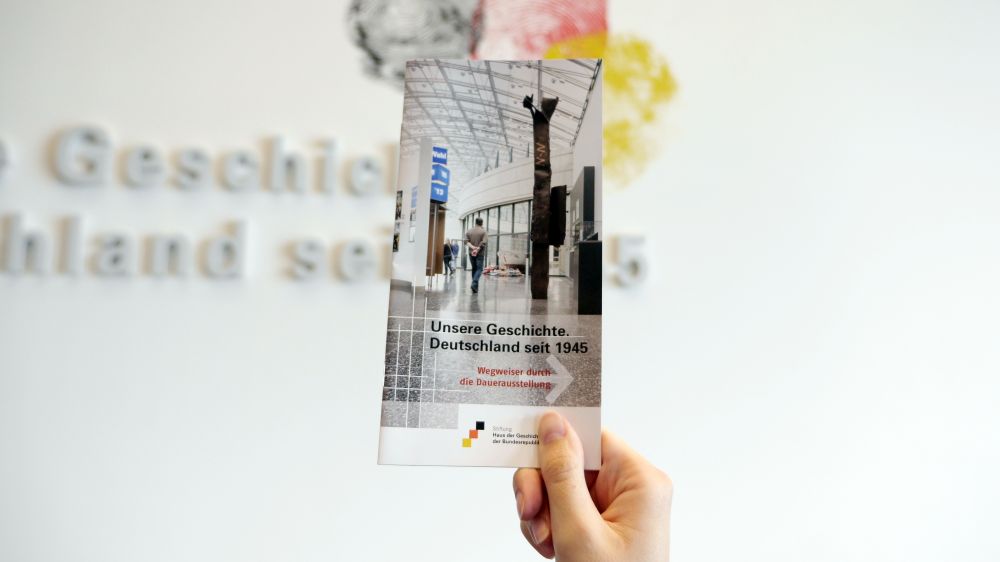 Eine Hand hält das Wegweiser-Heft vor der Dauerausstellung
