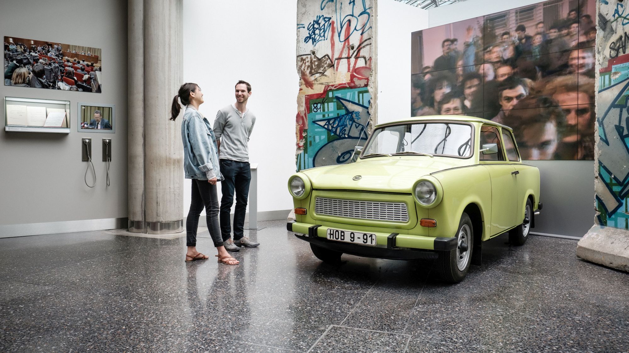 Eine junge Frau und ein junger Mann unterhalten sich in der Ausstellung neben einem grünen Trabi, der zwischen zwei großen Mauerteilen der Berliner Mauer steht.