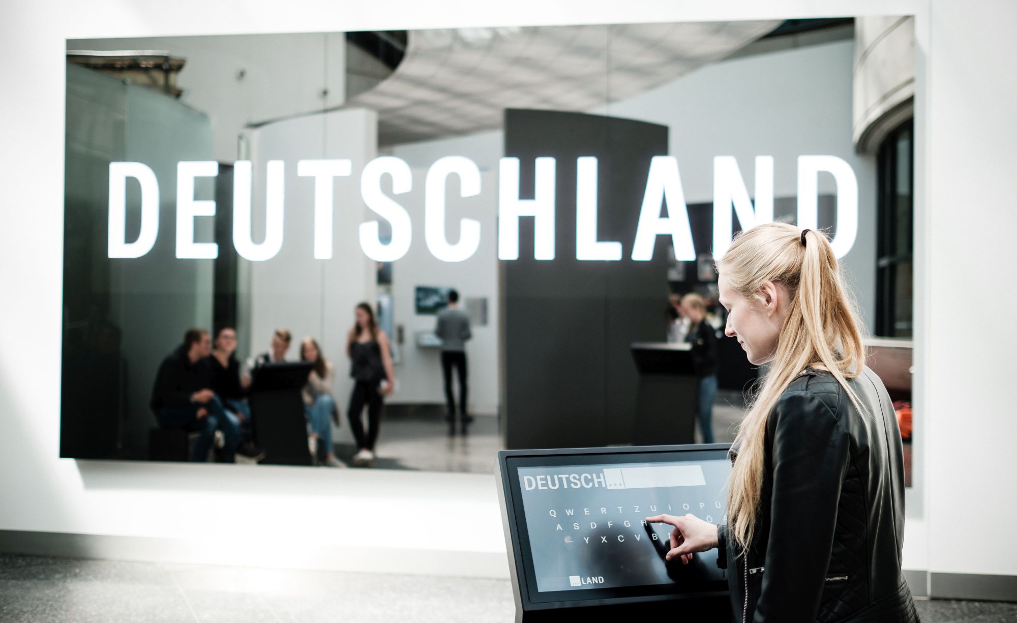 Eine junge Frau steht seitlich mit ihrem Finger auf einem Monitor mit Tastatur. Das Wort Deutschland wird groß auf eine spiegelnde dunkle Fläche geworfen, die ihr gegenüber an der Wand hängt.