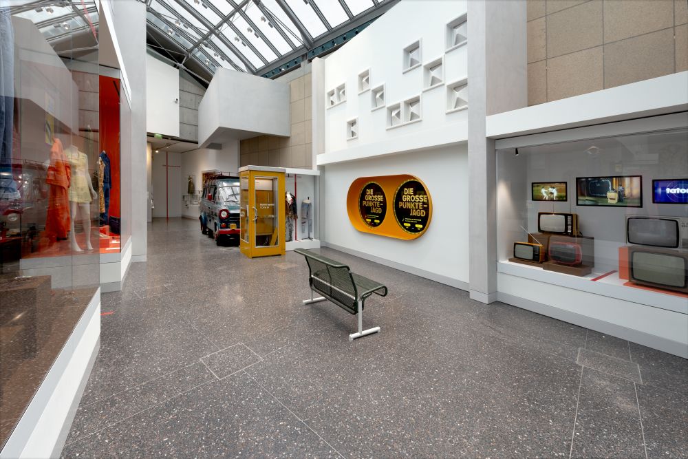 Schaufenster mit Kleidung, eine Telefonzelle und eine Bank vermitteln den Eindruck einer Fußgängerzone, im Hintergrund ein Ford Transit