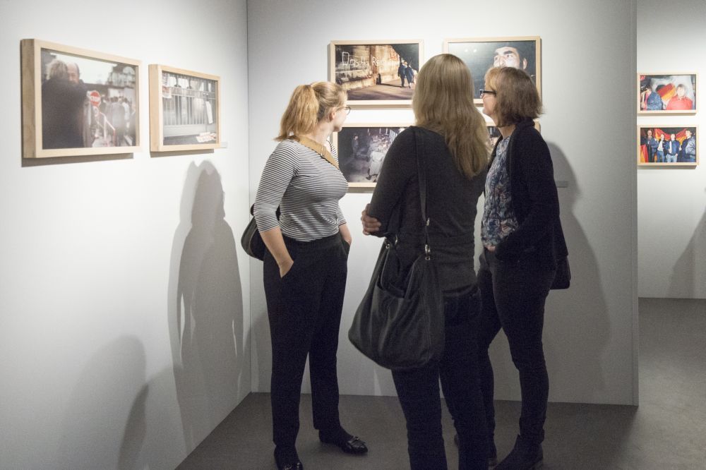 Drei junge Frauen betrachten gerahmte Fotografin an einer Zwischenwand, weitere Bilder hängen um sie herum