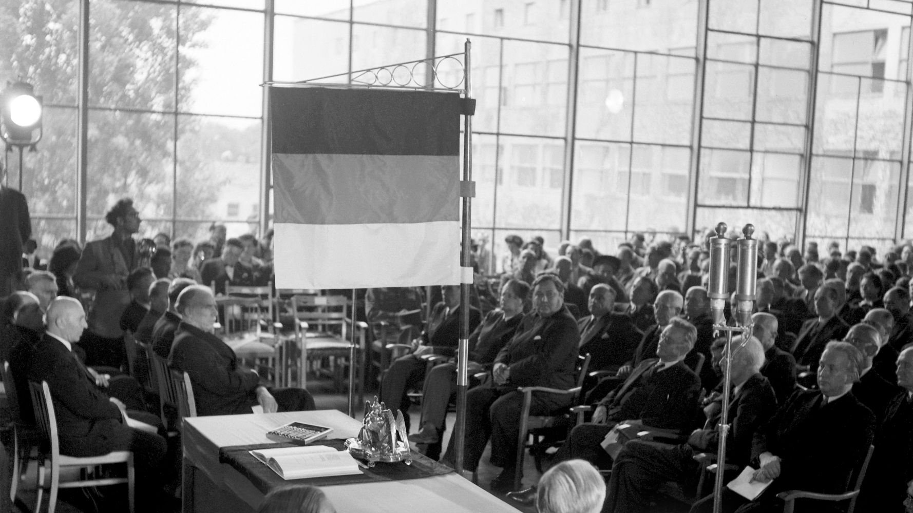 In einem vollen Saal mit großer Fensterfront sitzen viele Männer in Anzügen in Stuhlreihen. Im Vordergrund ist ein Tisch mit einer aufgeschlagenen Ausgabe des Grundgesetzes zu sehen, darüber steht eine Deutschlandflagge.