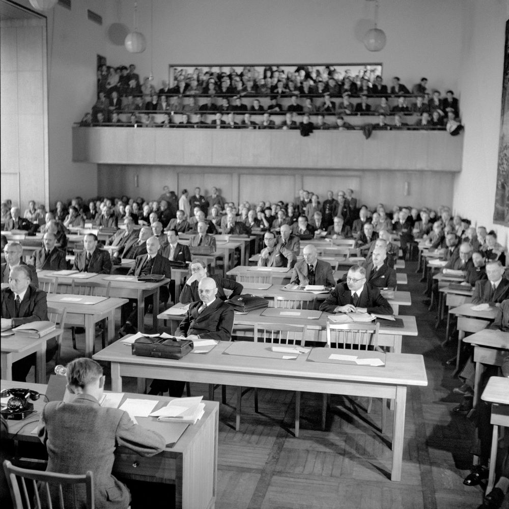 Blick ins Plenum und auf die Zuschauertribüne in der Pädagogischen Akademie. Im Plenum stehen Dreiertische, auf der Tribüne gibt es Sitzreihen.