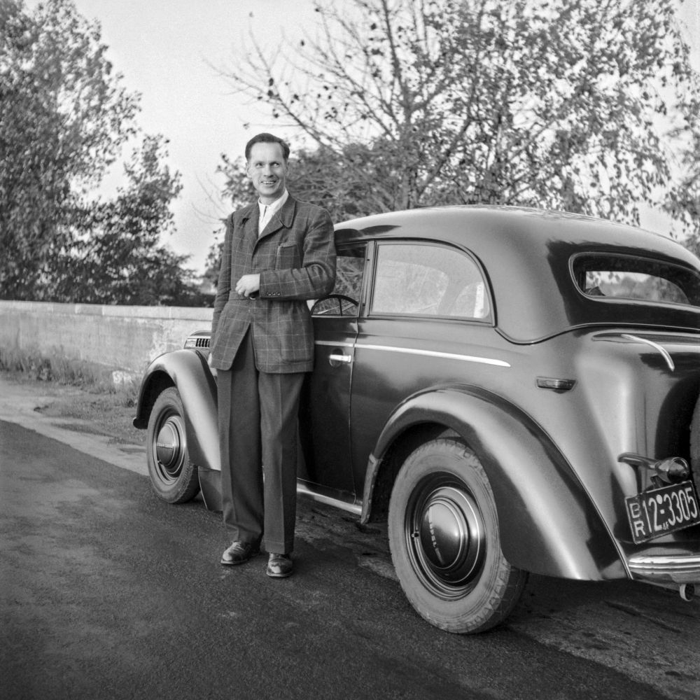 Ein junger Mann in Blazer und Stoffhose steht neben einem dunklen VW Käfer.