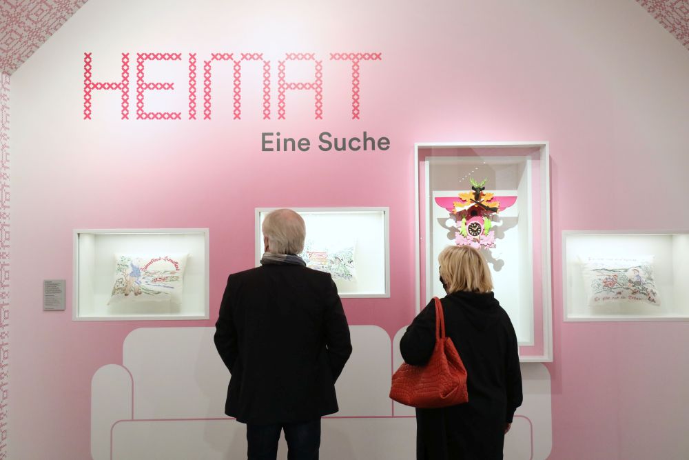 Ein älteres Paar steht vor einer Ausstellungwand mit eingelassenen Vitrinen und einem aufgemalten Sofa.