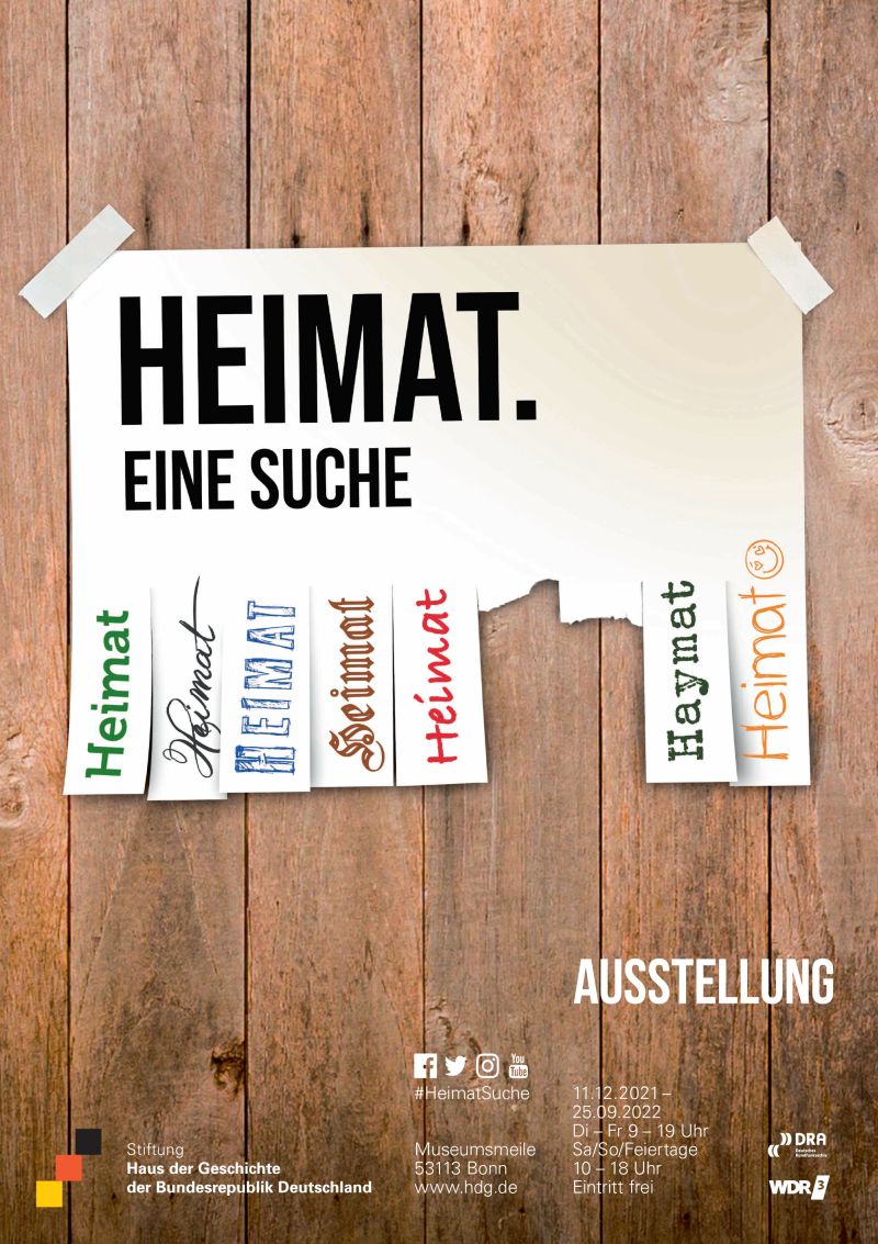 An einer Holzwand hängt ein Abreisszettel, auf dem in vielen verschiedenen Farben und Schriftarten das Wort Heimat steht.