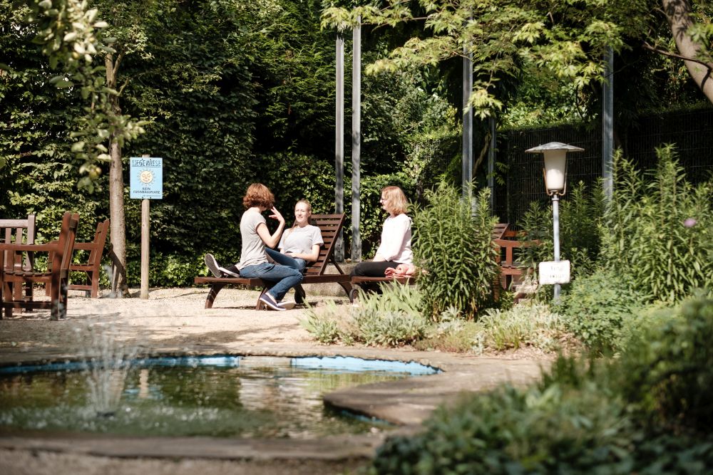 Drei junge Frau sitzen im Bildhintergrund auf Liegestühlen zusammen. Im Vordergrund gibt es einen Teich mit einem kleinen Springbrunnen, rundherum grüne Büsche und Bäume.