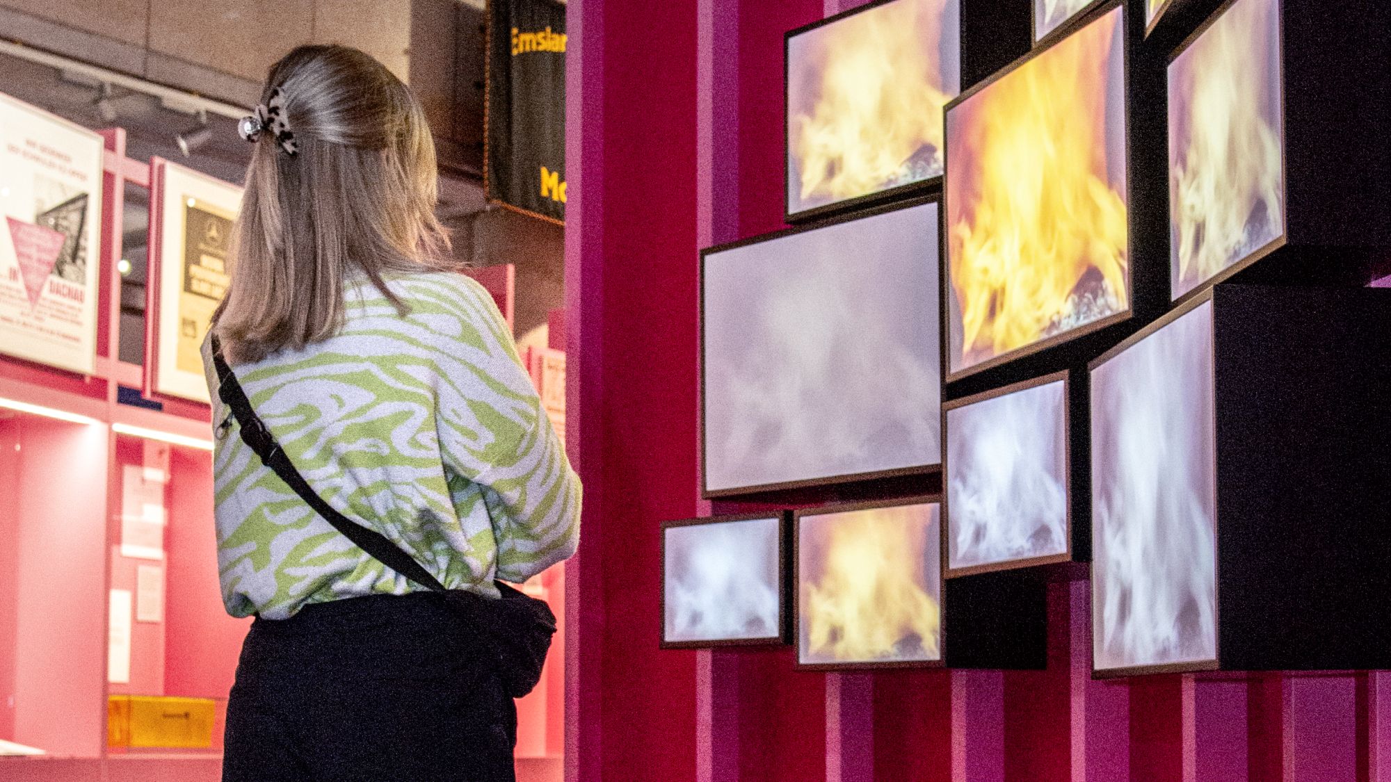 Eine blonde Frau, die von schräg hinten zu sehen ist, schaut in einem Ausstellungsraum auf Monitore, auf denen Flammen zu sehen sind.
