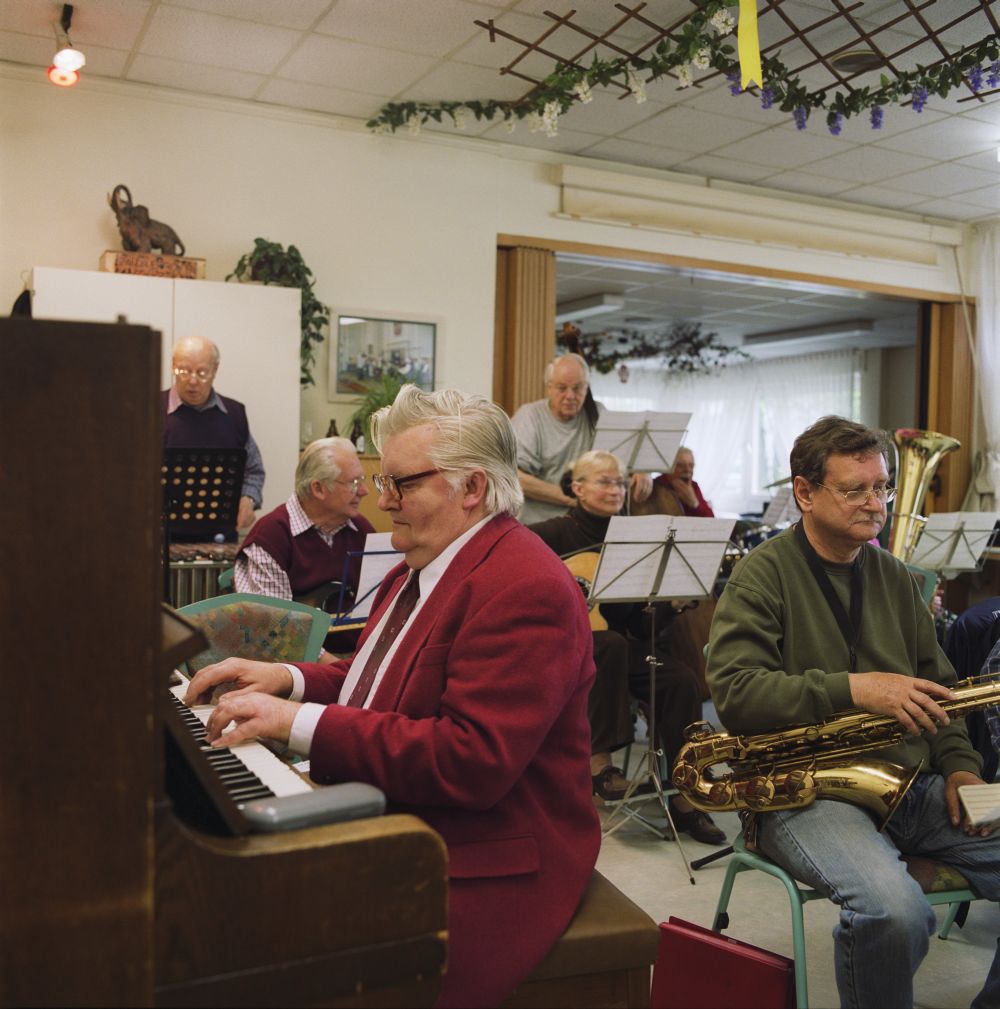 Ein älterer Mann sitzt in einem Zimmer am Klavier, hinter ihm weitere Senioren mit Instrumenten und Notenständern