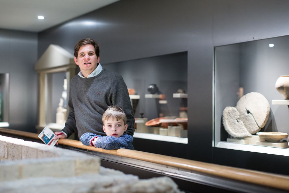 Ein kleiner junge und ein Mann stehen an einem Geländer. Hinter ihnen sind große Schaukästen in einer Wand zu sehen mit Vasen und einem zerbrochenen Mühlstein