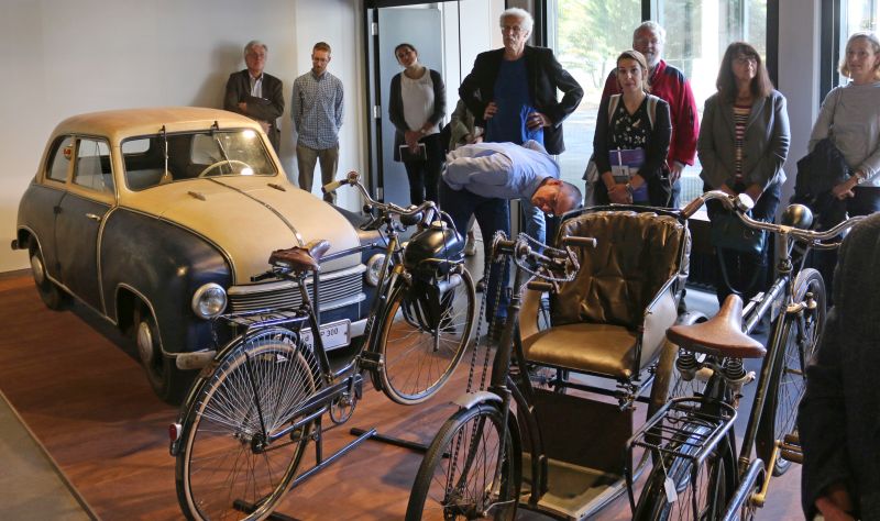 A group of people of different ages is standing next to a car, in the foreground are bicycles of different models.