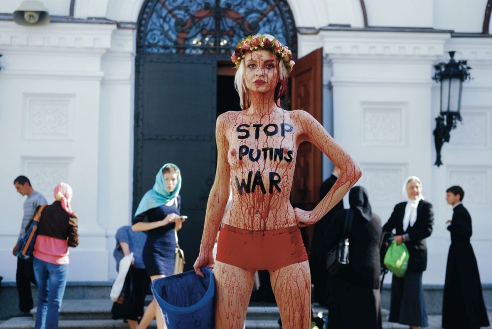 Eine Aktivistin der Frauenbewegung FEMEN protestiert gegen den Einmarsch russischer Truppen in die Ukraine. Auf ihrem freien Oberkörper ist "Stop Putins War" geschrieben, ihr ganzer Körper ist mit einer roten Flüssigkeit übergossen, auf dem Kopf trägt sie einen Blumenkranz.