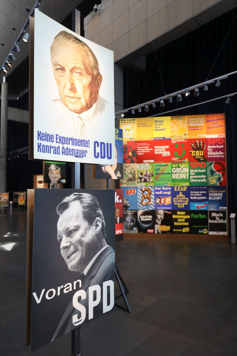 Das berühmte CDU-Plakat mit Konrad Adenauer und dem Slogan Keine Experimente sowie Willy Brandt und der Slogan Voran SPD im Vordergrund und eine große Wand mit vielen weiteren Plakaten im Hintergrund.