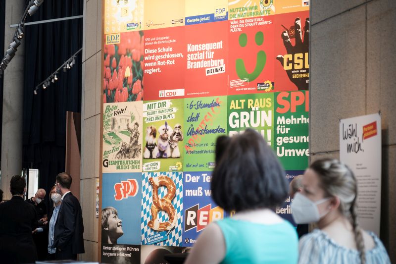 Zwei Frauen vor einer bunten Wand aus nach Farbe angeordneten Wahlplakaten.