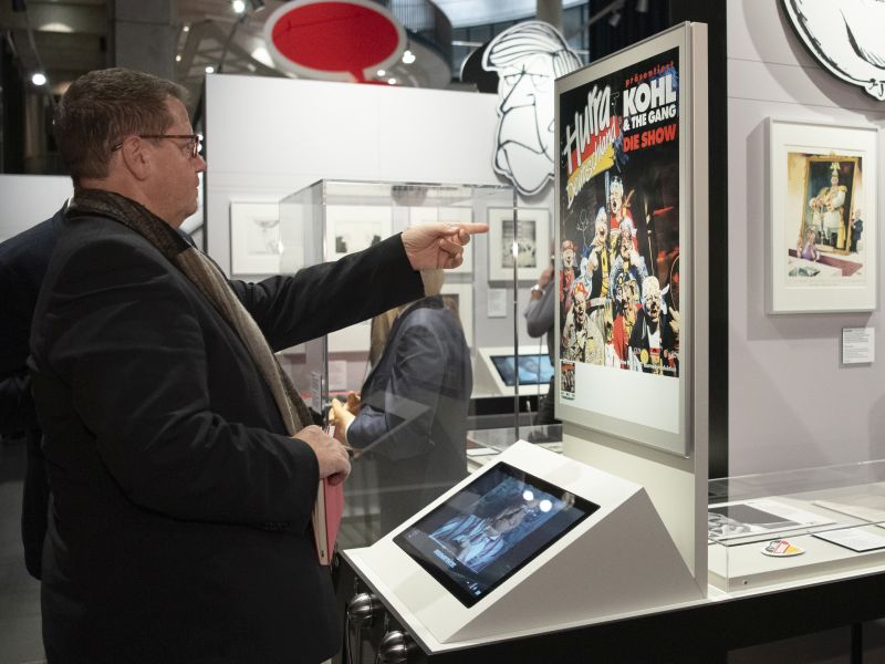 Besucher vor einem Bildschirm und einem satirischen Plakat zu Helmut Kohl.