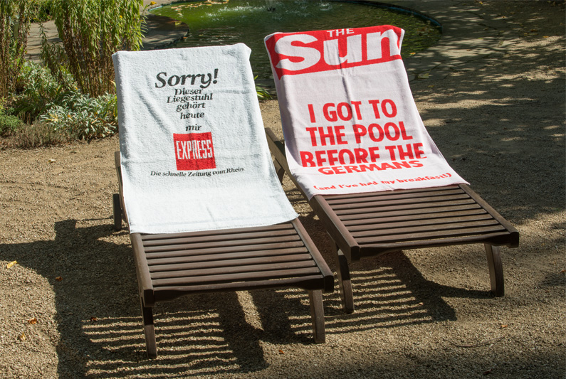 Zwei Liegestühle mit Handtüchern, eines vom Express, "Sorry, dieser LIegestuhl gehört heute mir!" und eines von der britischen Boulevardzeitung "The Sun", "I got to the pool before the Germans".