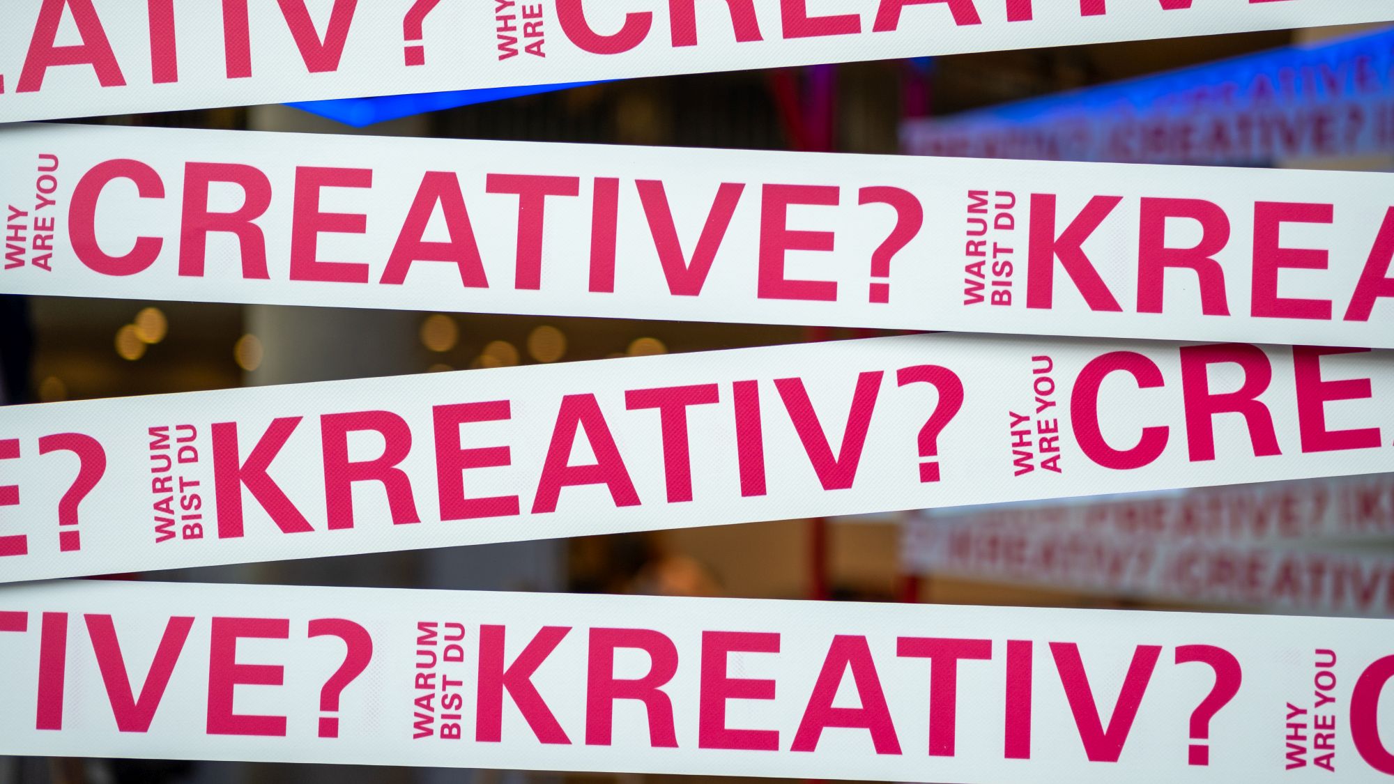 Das Foto zeigt Nahaufnahmen weißer Bänder mit pinkfarbener Schrift und der deutsch-englischen Aufschrift Warum bist du kreativ bzw. Why are you creative?