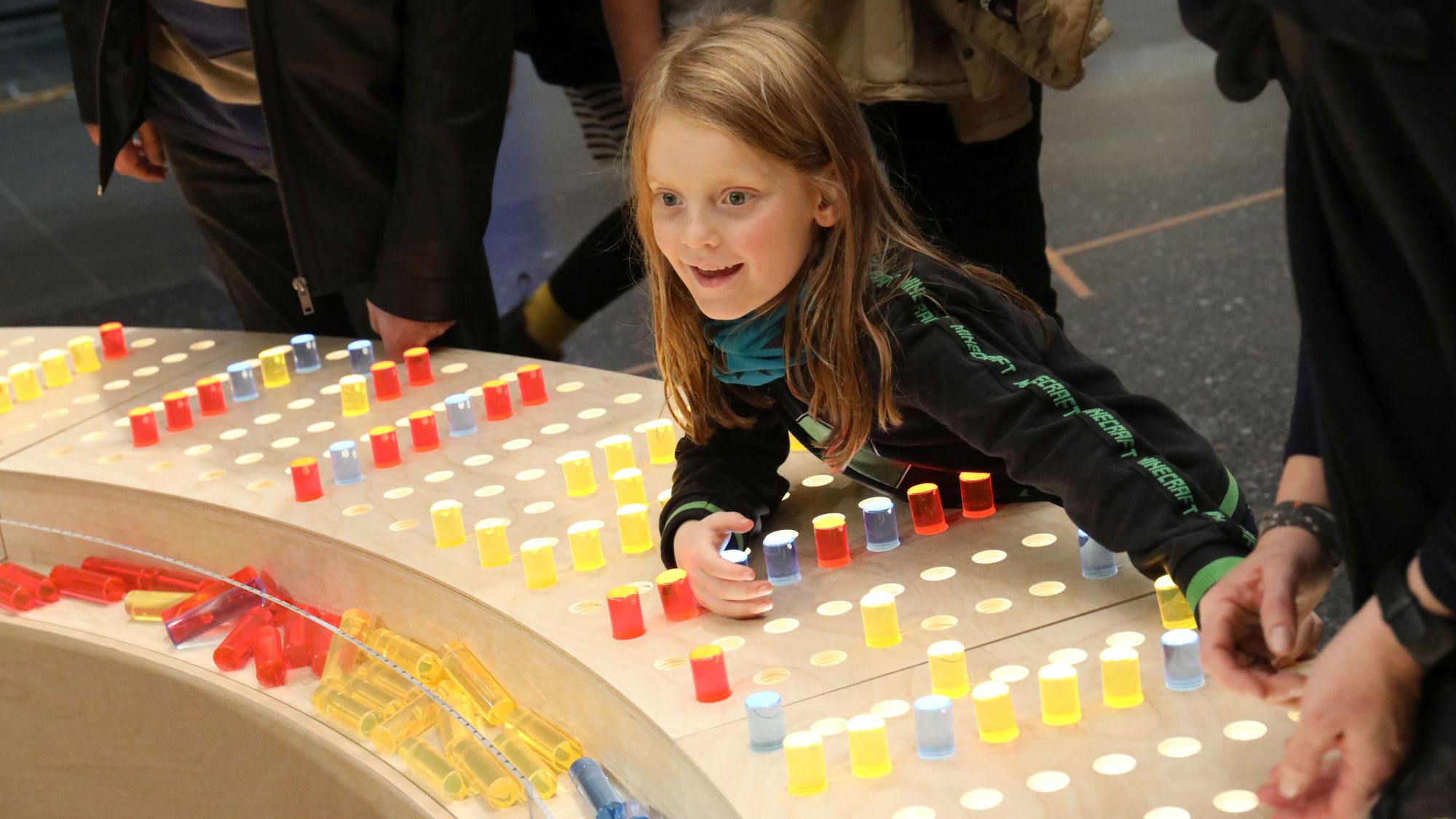 Ein etwa fünfjähriges Mädchen lehnt mit begeistertem Gesichtsausdruck über einer Holztheke mit bunten Steckelementen.