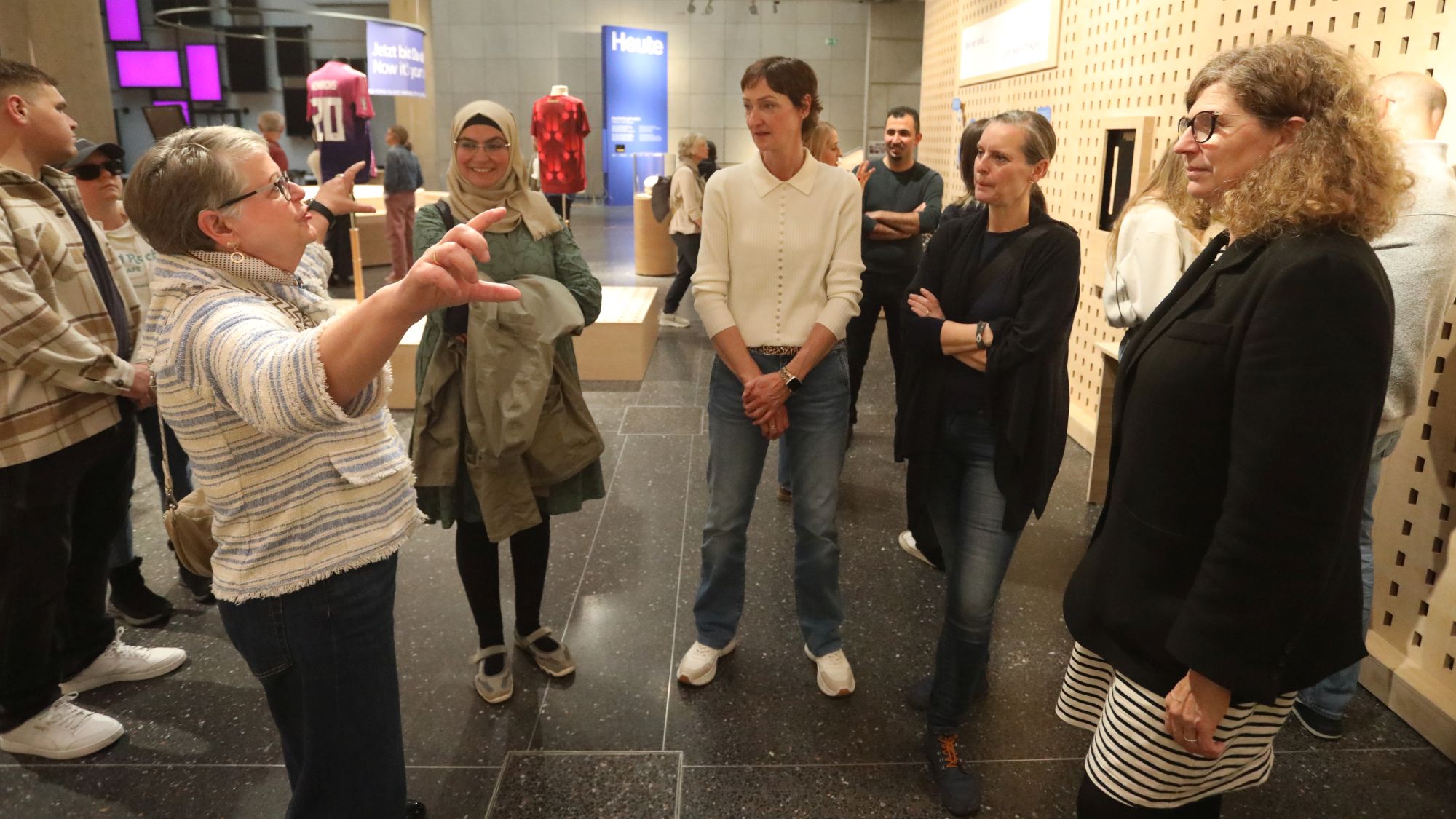 Vier Frauen stehen im Halbkreis eine fünfte Frau herum, die zu ihnen spricht und mit erhobenen Armen gestikuliert. 