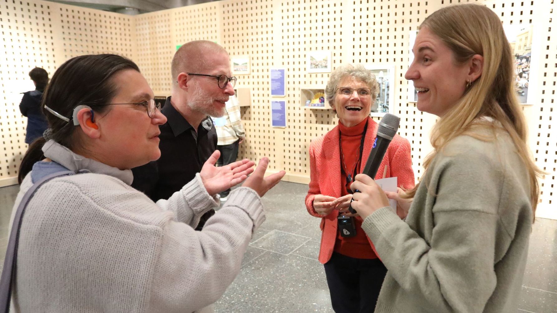 Eine Frau mit Mikrofon steht mit zwei anderen Frauen und einem Mann in einem kleinen Kreis. Die drei sind ihr zugewandt. Eine Frau spricht mit ihr und hebt gestikulierend die Hände, die andere lacht sie fröhlich an.