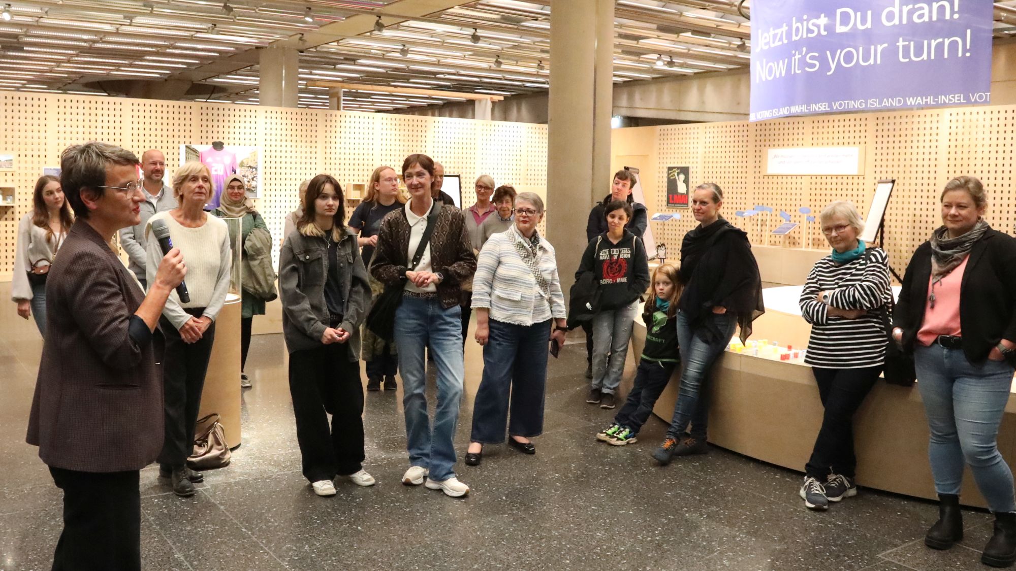 Im Vordergrund eine Frau, die in ein Mikrofon spricht. Daneben Frauen und Männer unterschiedlichen Alters, die ihr zuhören. 