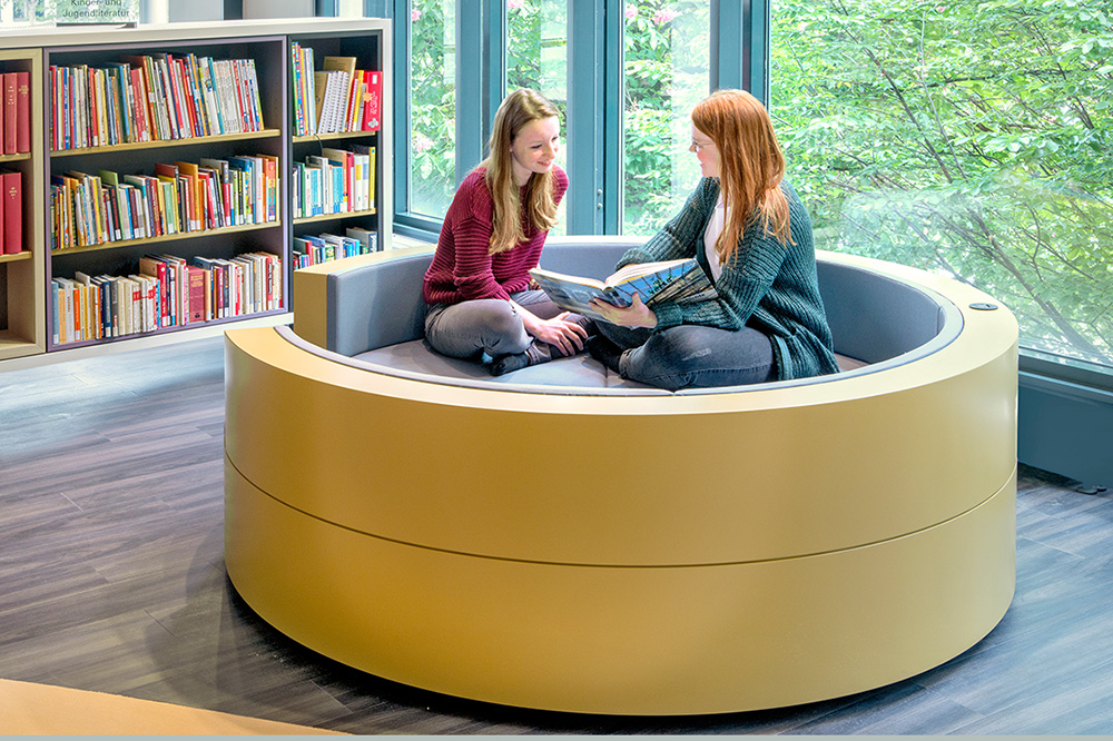 Zwei junge Frauen sitzen auf einem Sofa in der Bibliothek, dahinter steht ein Bücherregal.