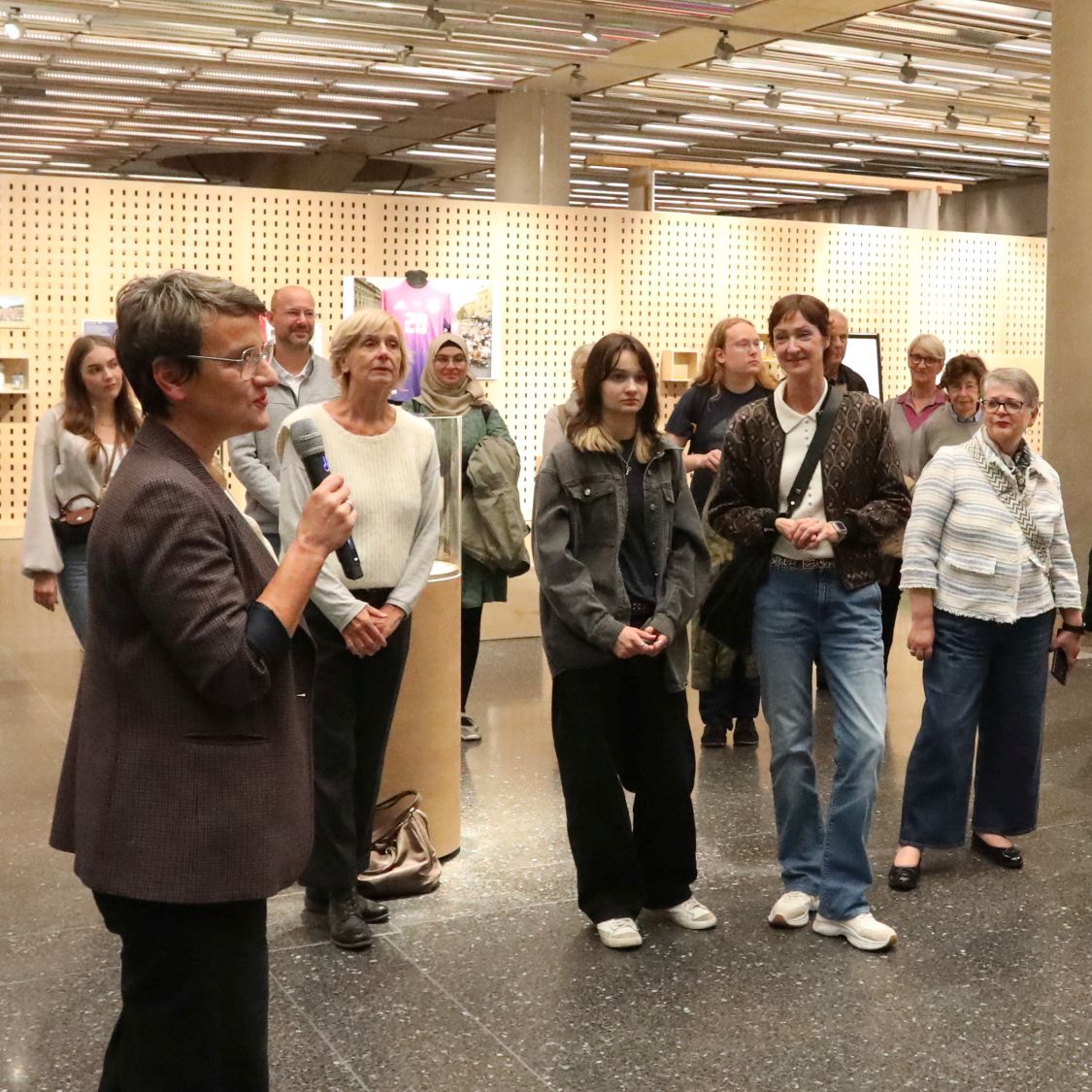 Eine Begleitung in der Ausstellung Im Vordergrund eine Frau, die in ein Mikrofon spricht. Im Hintergrund Frauen und Männer, die ihr zuhören.
