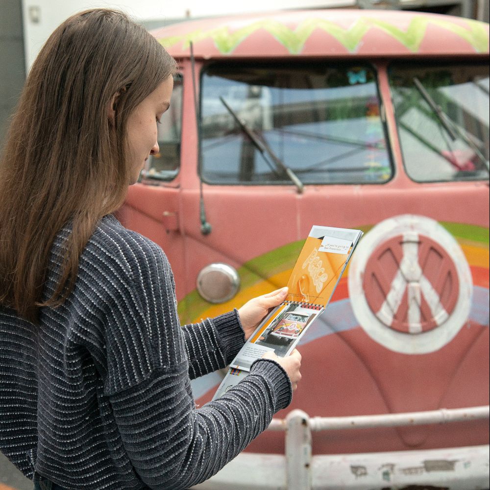 Junge Frau mit einem Block in der Hand. Im Hintergrund befindet ein rosa VW-Bus aus der Dauerausstellung.