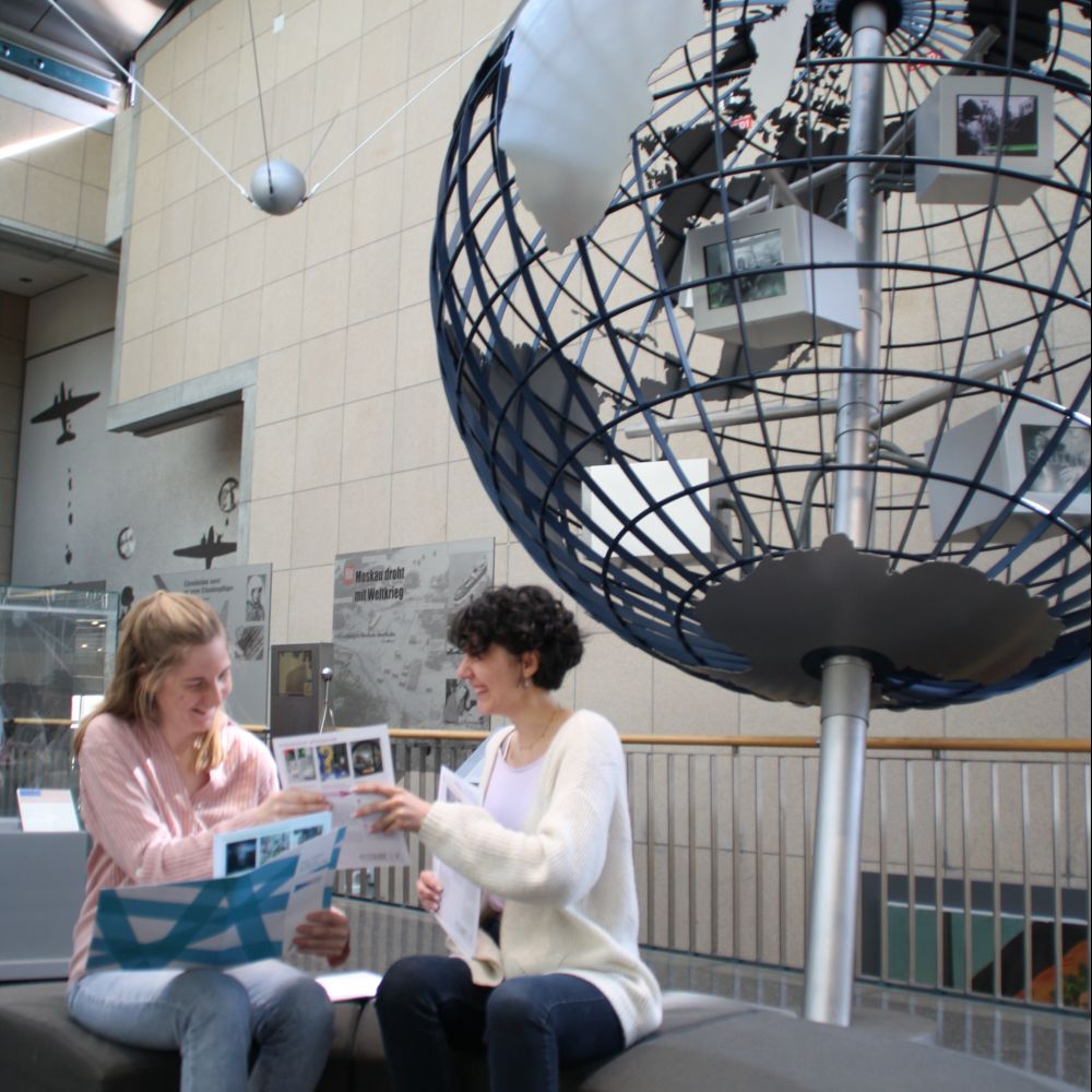 Zwei junge Frauen mit der Materialmappe. Im Hintergrund befindet sich eine Weltkugel mit Monitoren.