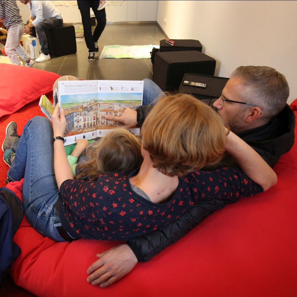 Familie auf einem roten Sitzkissen. Die drei Personen halten ein Bilderbuch in der Hand.