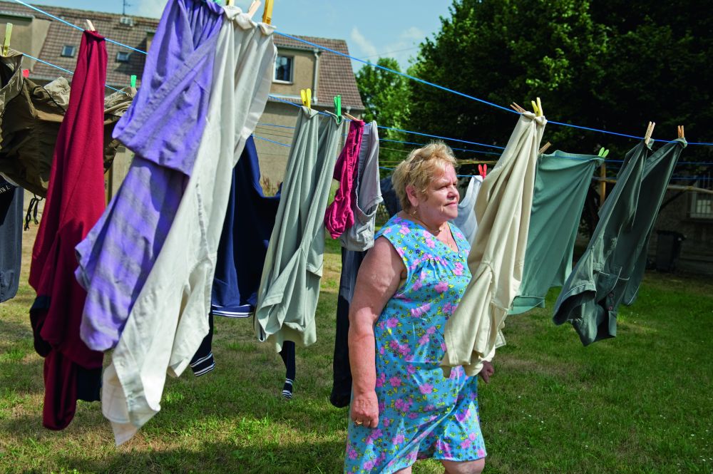 Ältere Frau steht auf einer grünen Wiese inmitten von Wäscheleinen, an denen bunte Kleidungsstücke hängen.