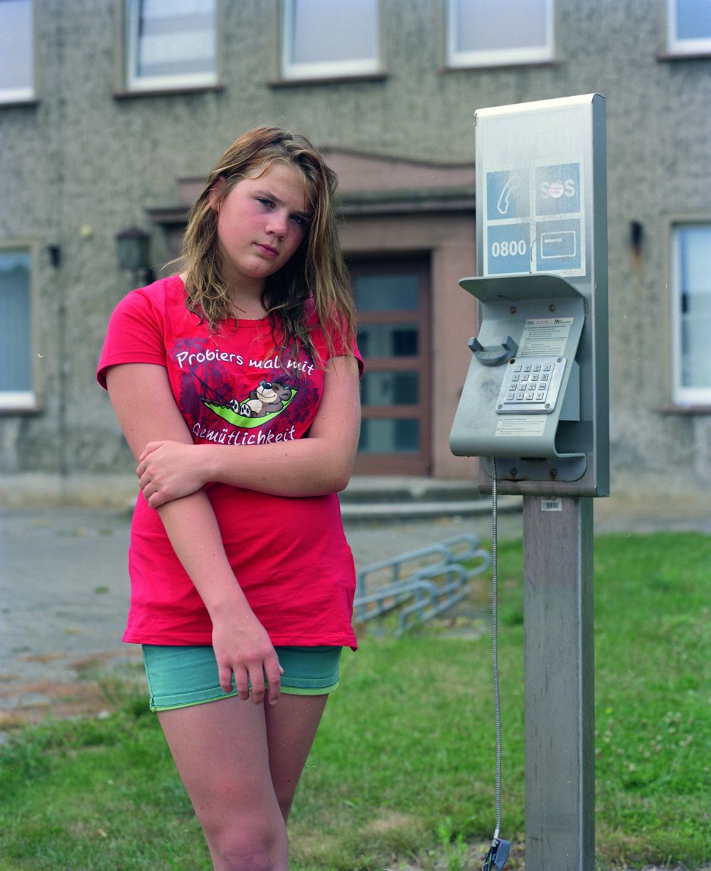 Ein Mädchen steht vor einem grauen Haus, neben ihr befindet sich eine sich offensichtlich nicht in Betrieb befindende Telefonstation.