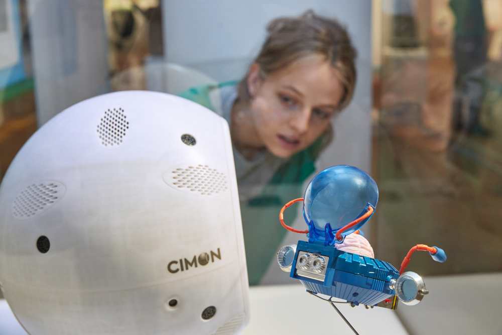 A young woman looks at Cimon, the world's first astronaut assistant and the toy figurine "Professor Simon Wright" from the animated series "Captain Future".