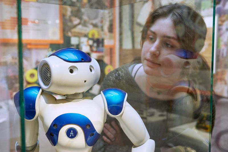 A young woman looks at a robot which is displayed behind glass
