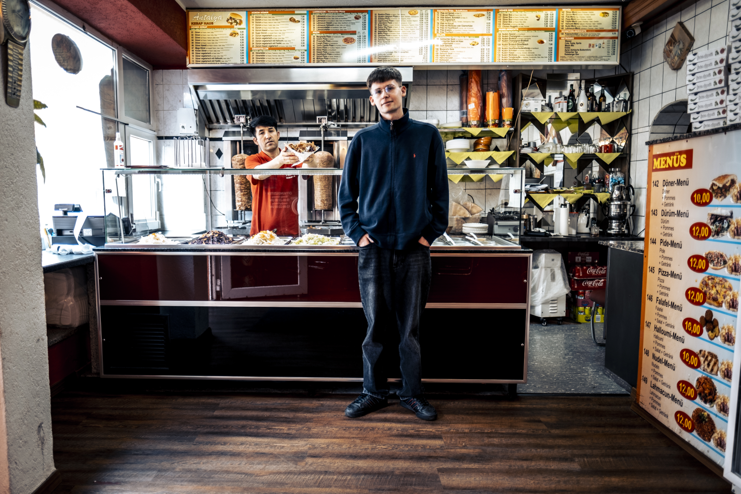 Ein junger Mann steht in einem Kebab-Imbiss. Hinter ihm serviert ein Mitarbeiter des Imbisses gerade einen Döner. 