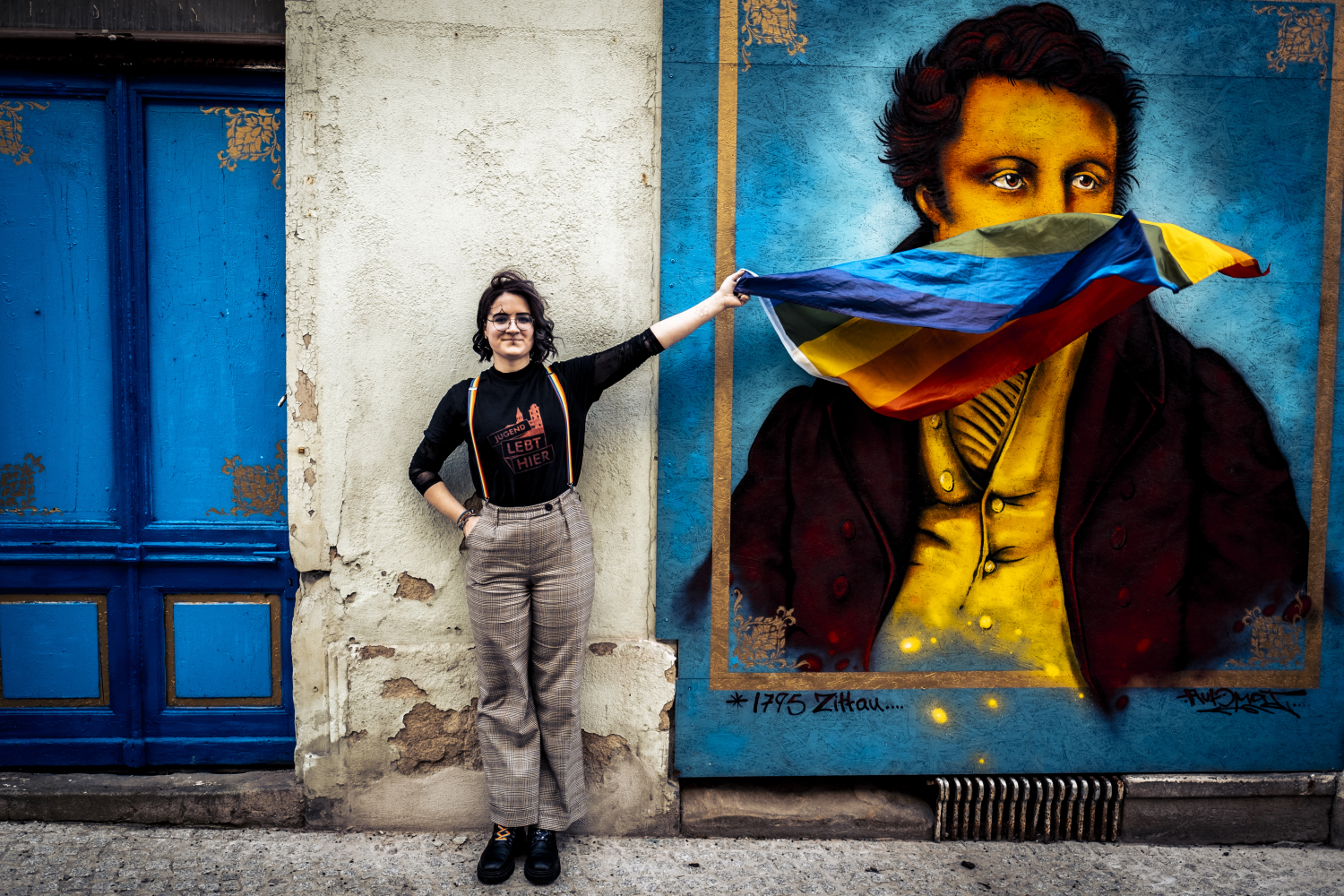 Eine junge Frau mit Regenbogenflagge in der Hand steht vor einer Wand. Auf der Wand ist ein Mann in Kleidung des 18. Jahrhunderts abgebildet. Die Frau lässt die Flagge über sein Gesicht wehen.