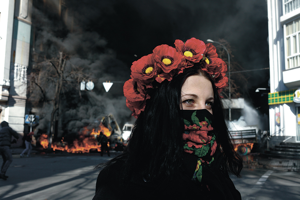 Junge Frau mit Blumenkranz auf Kopf steht auf Straße. Im Hintergrund brennende Autos und schwarzer Rauch.