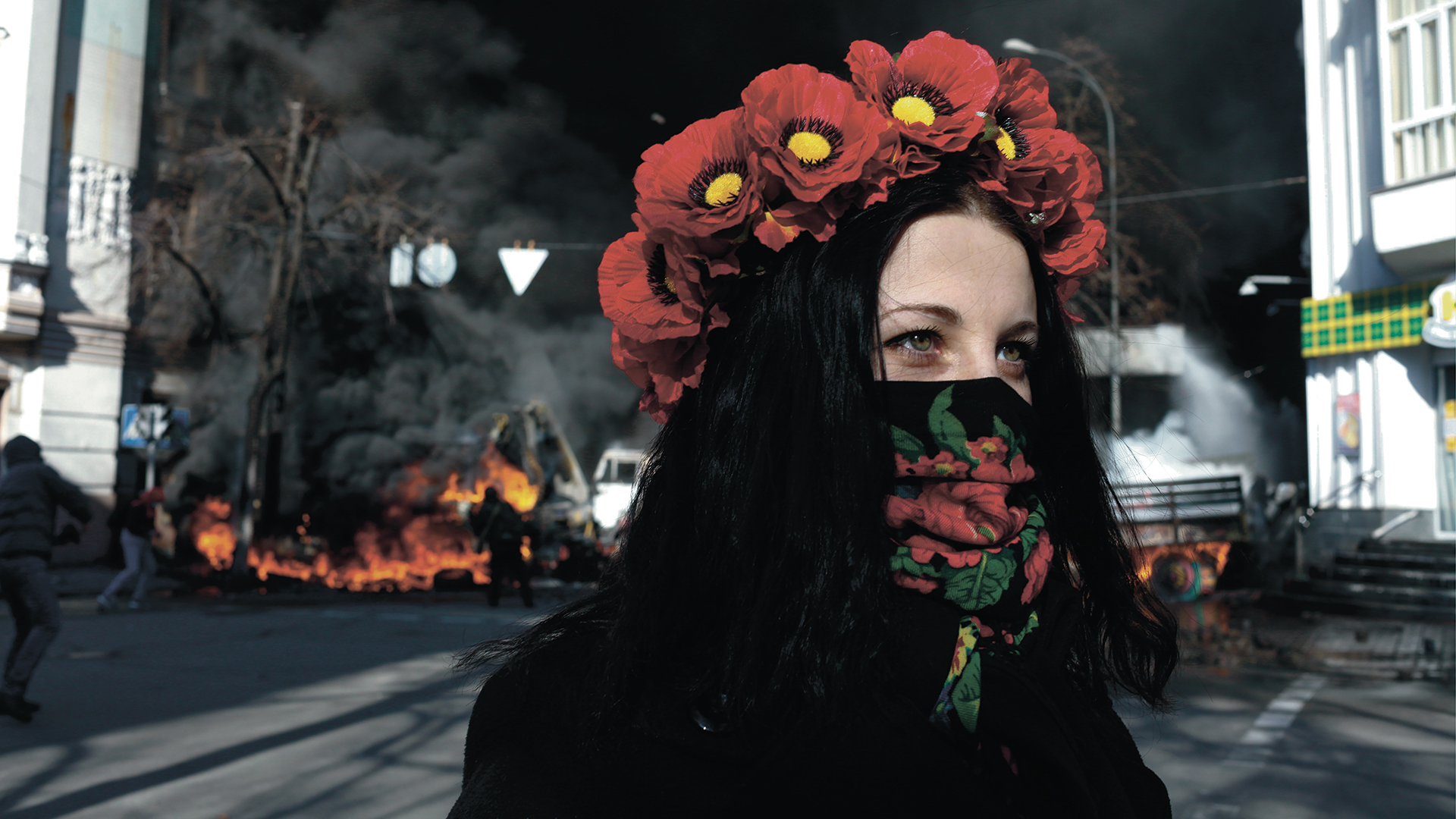 Junge Frau mit Blumenkranz auf Kopf steht auf Straße. Im Hintergrund brennende Autos und schwarzer Rauch.