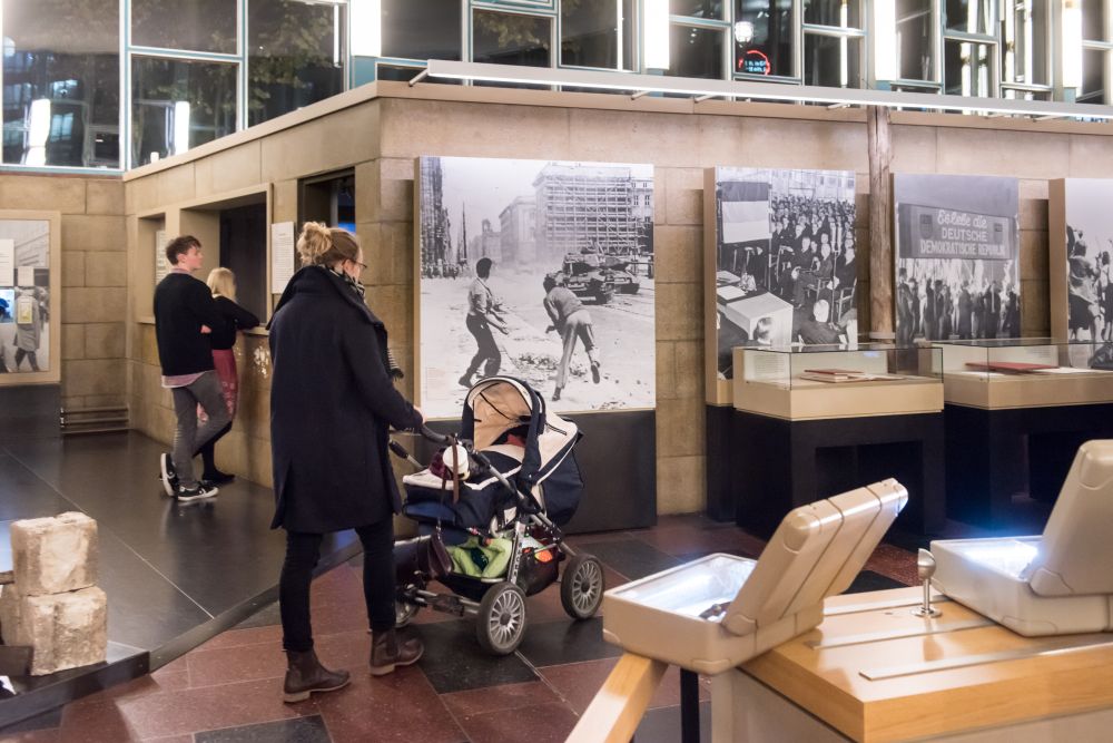 Eine junge Frau geht mit einem Kinderwagen in die Ausstellung, es sind großformatige Fotos und geöffnete Koffer zu sehen