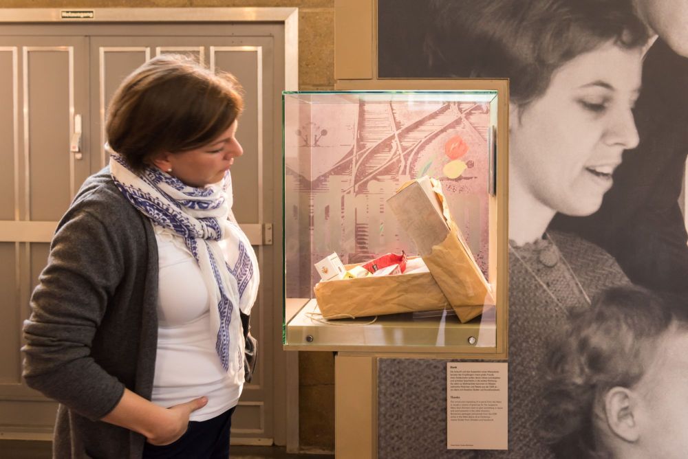 Eine Frau steht seitlich vpr einer Vitrine mit einem geöffnete Paket, darin verschiedene Westdeutsche Produkte, im Hintergrund ein großformatiges Fotos mit einer jungen Frau