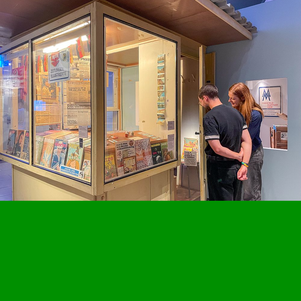 Eine Begleiterin zeigt einem Besucher ein Objekt an einem Zeitungskiosk in der Ausstellung