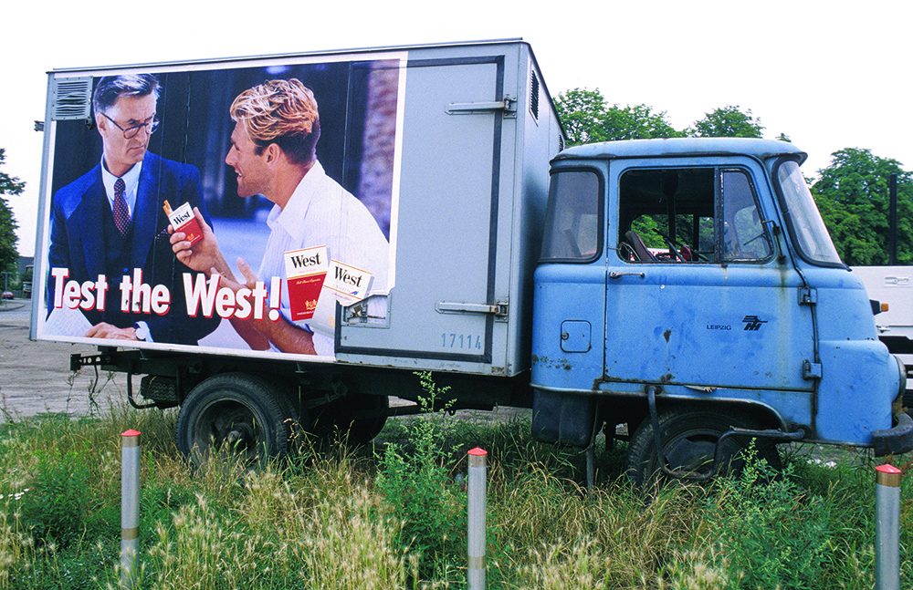 Ein kleiner LKW mit Werbung für Zigaretten. Auf dem Werbeplakat bietet ein junger Mann einem Geschäftsmann eine Zigarette an.