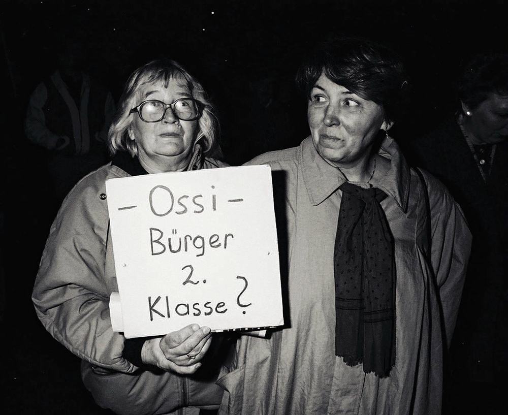 Fotografie in schwarz-weiß. Darauf sind zwei Frauen zu sehen. Die linke hält ein Plakat hoch, auf dem steht: Ossi. Bürger zweiter Klasse? Die rechte Frau schaut die linke Frau an.