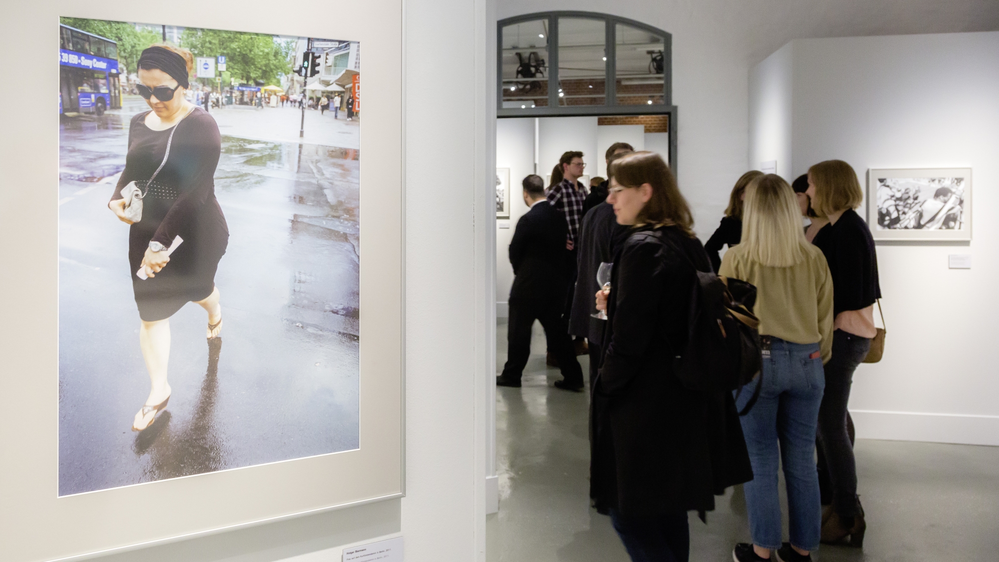 Besucher in einem Ausstellungsraum mit weißen Wänden, im Vordergrund eine Frau mit Kleid, Sonnenbrille und Handtasche.